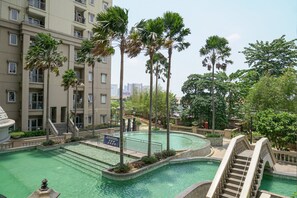 A swimming pool area with palm trees, a bridge, and a multi-story building in the background. - Homey 2BR Apartment at Grand Palace Kemayoran (Jakarta)