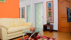 Un salon avec un canapé jaune, une table basse en verre, un tapis à motifs floraux rouges et une porte en bois.