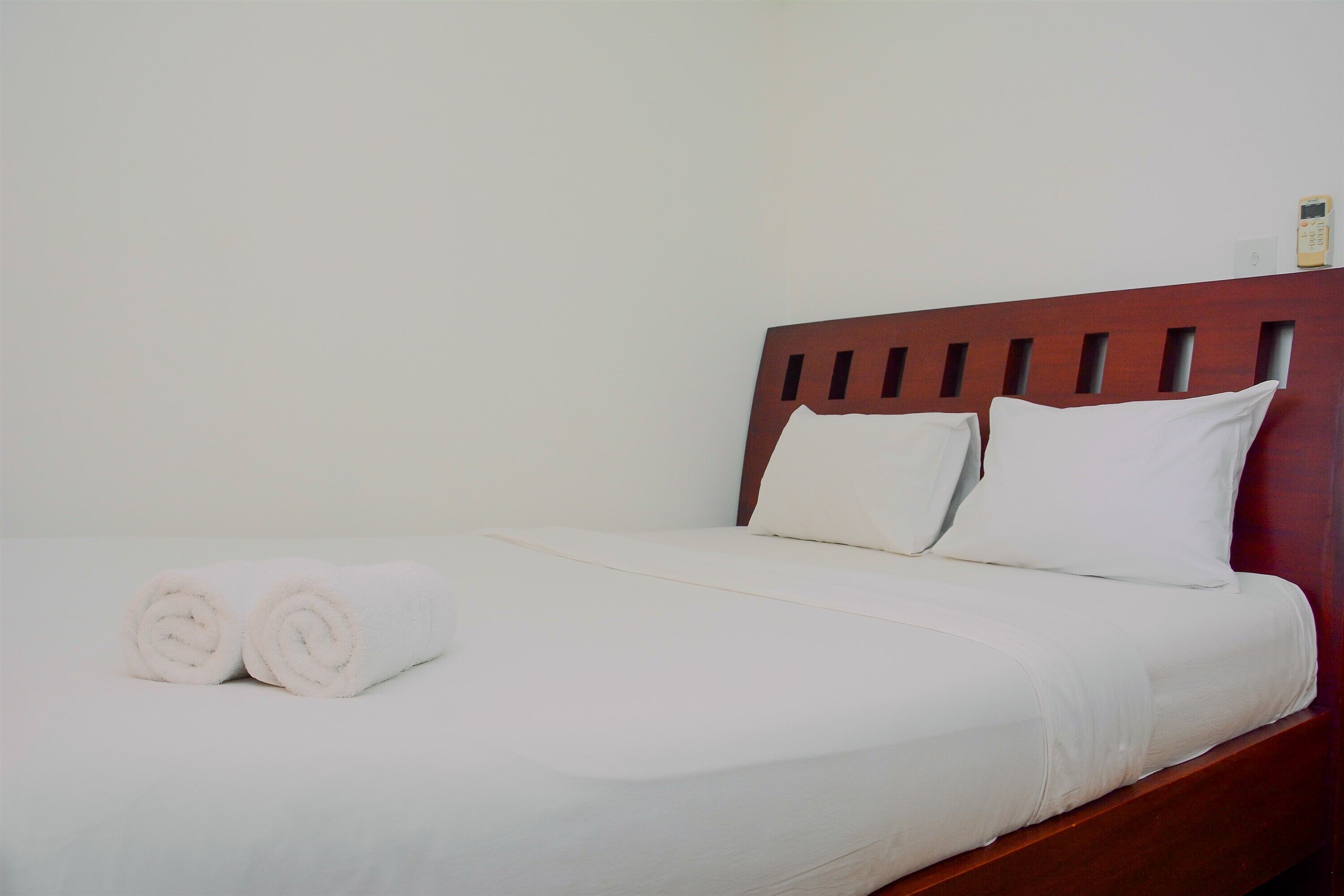 A neatly made bed with white linens and two rolled towels, a wooden headboard, and a wall-mounted thermostat.