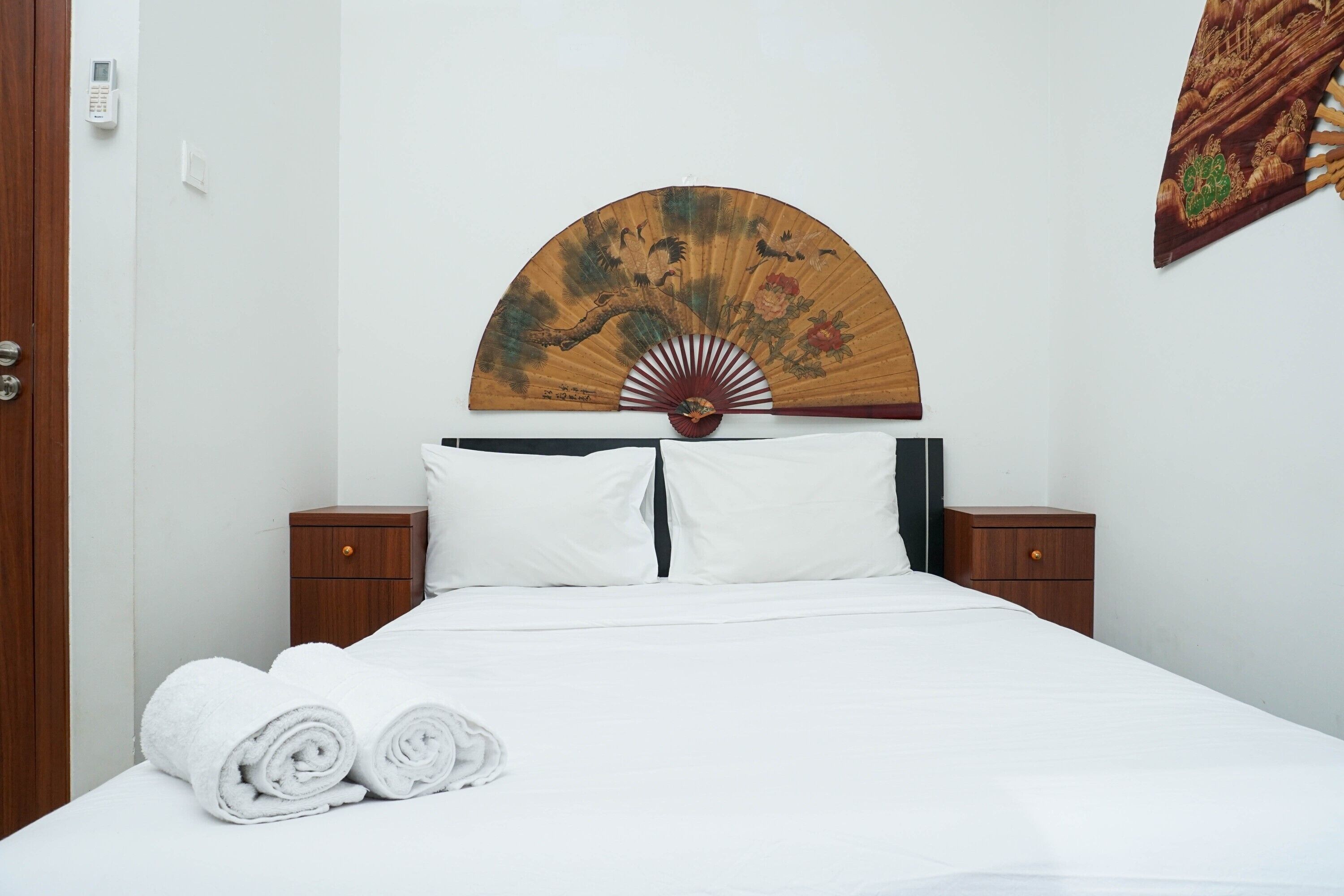 A neatly made bed with white linens, two wooden nightstands, a wall-mounted fan, and a folded white towel on the bed.