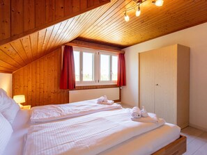 A bedroom with a wooden ceiling, a window with red curtains, a bed with white linens, and a wooden wardrobe.