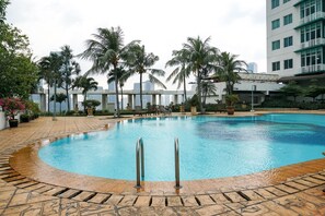 Una piscina con solárium de baldosas, palmeras y un edificio al fondo.