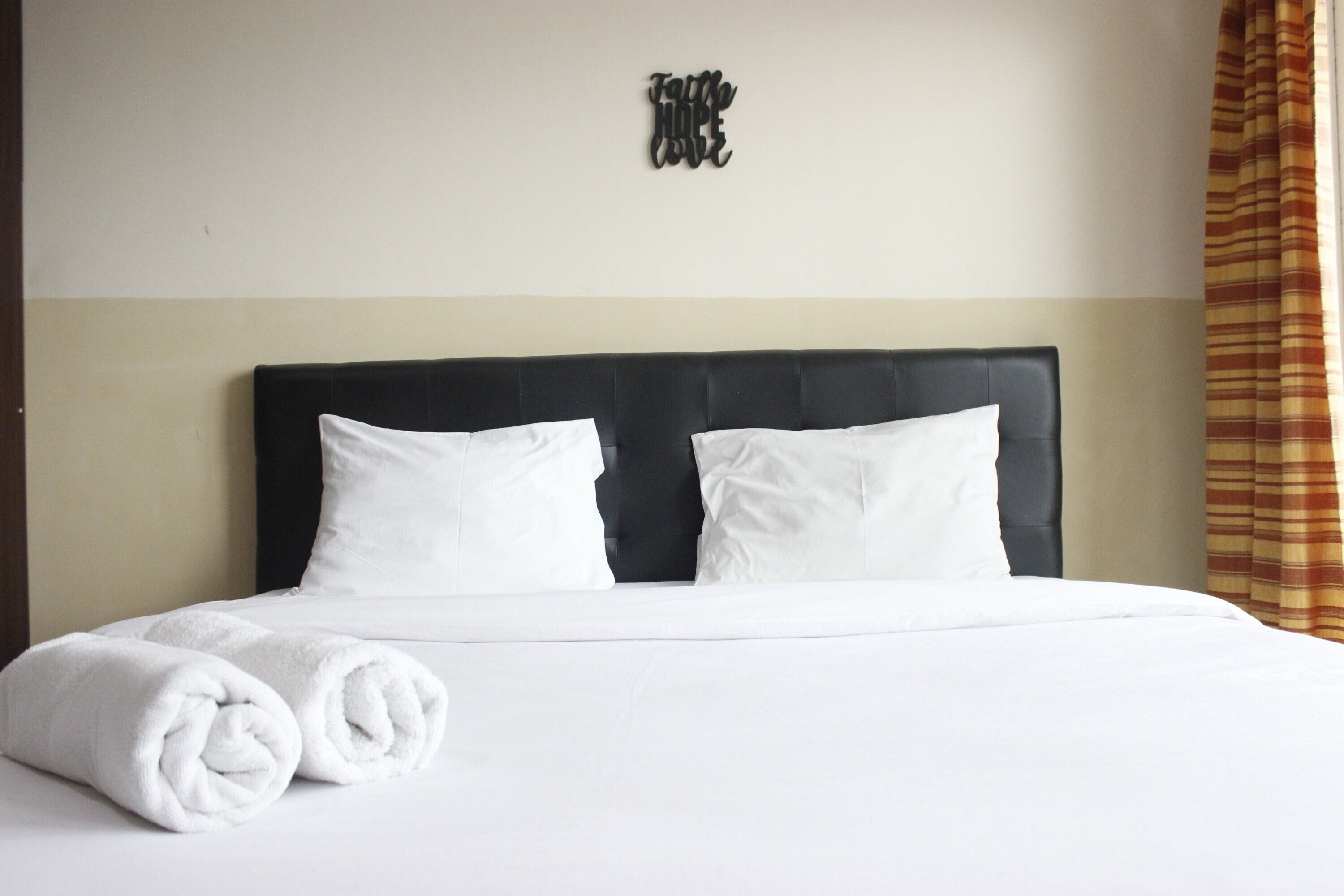 Una cama bien hecha con ropa de cama blanca y dos toallas enrolladas, un cabecero tapizado negro, y un letrero en la pared que dice 'Family Hope'.