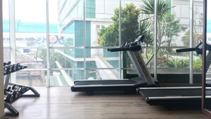 Une salle de sport équipée de tapis de course et d’haltères, avec de grandes fenêtres offrant une vue sur les bâtiments et la verdure.