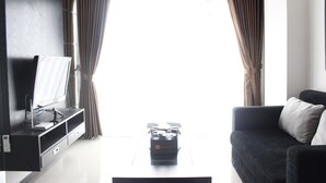 A modern living room with a flat-screen TV on a black entertainment unit, a dark sofa with a white pillow, and a travel suitcase on a black ottoman. - Comfortable & Gorgeous 2BR at Gateway Pasteur Apartment (Cimahi)