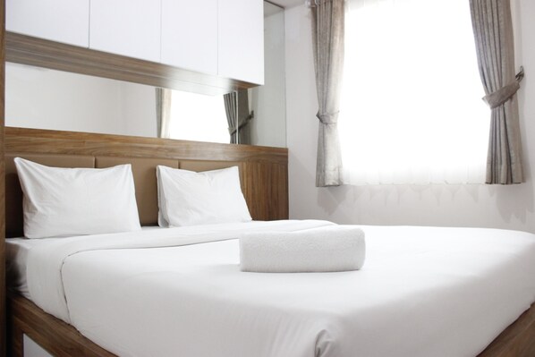 Una cama bien hecha con sábanas blancas y una toalla doblada, un cabecero de madera y una ventana con cortinas.