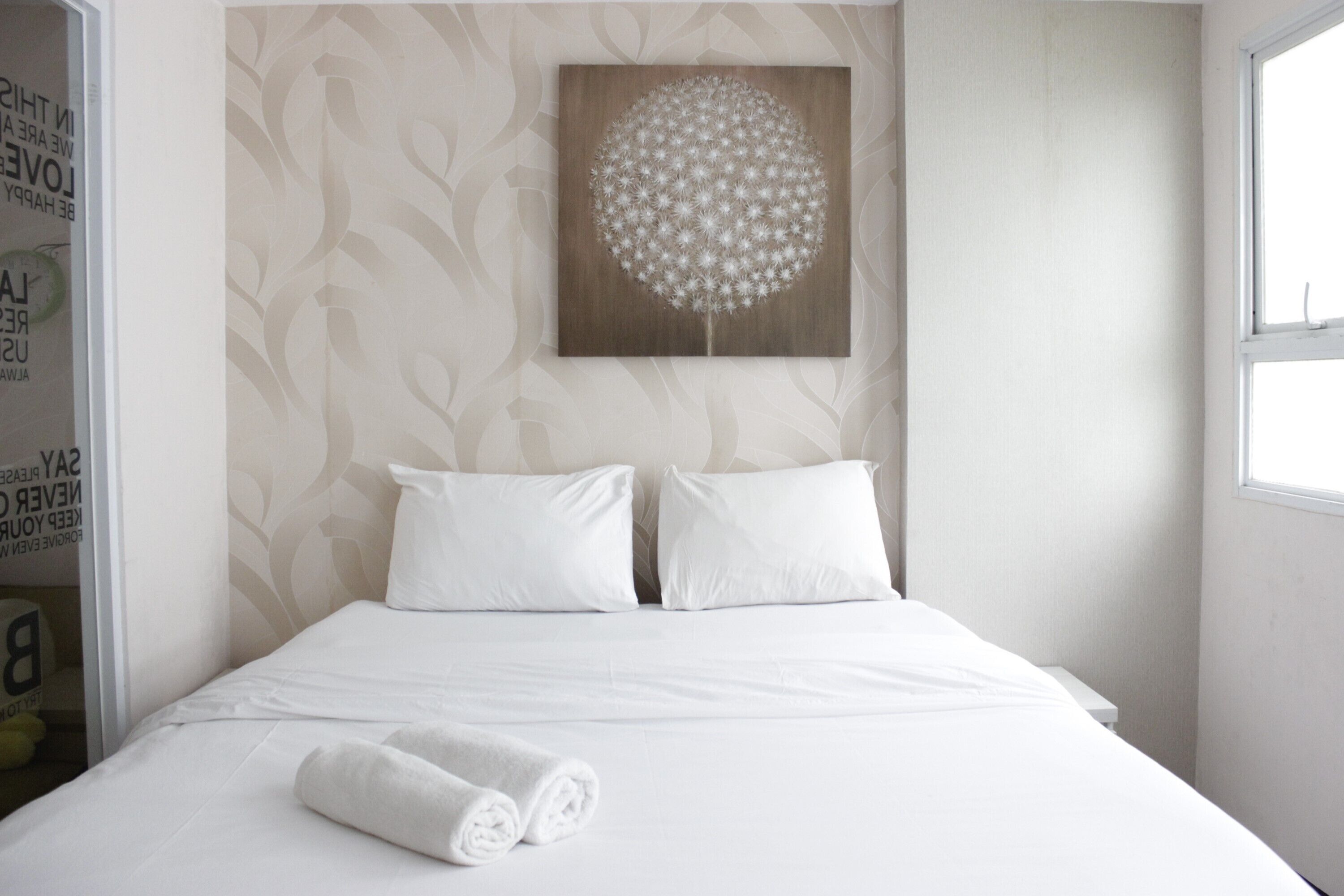 Cama bem arrumada com lençóis brancos e duas almofadas, uma obra de arte na parede retratando um dente-de-leão, e uma janela que oferece luz natural.