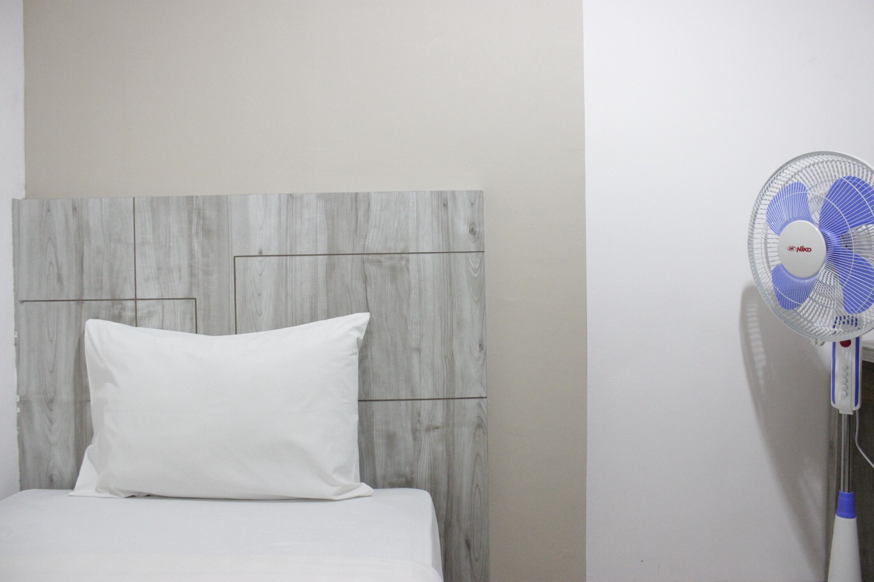 A bed with a white pillow and a wooden headboard. A standing fan is positioned to the right of the bed.