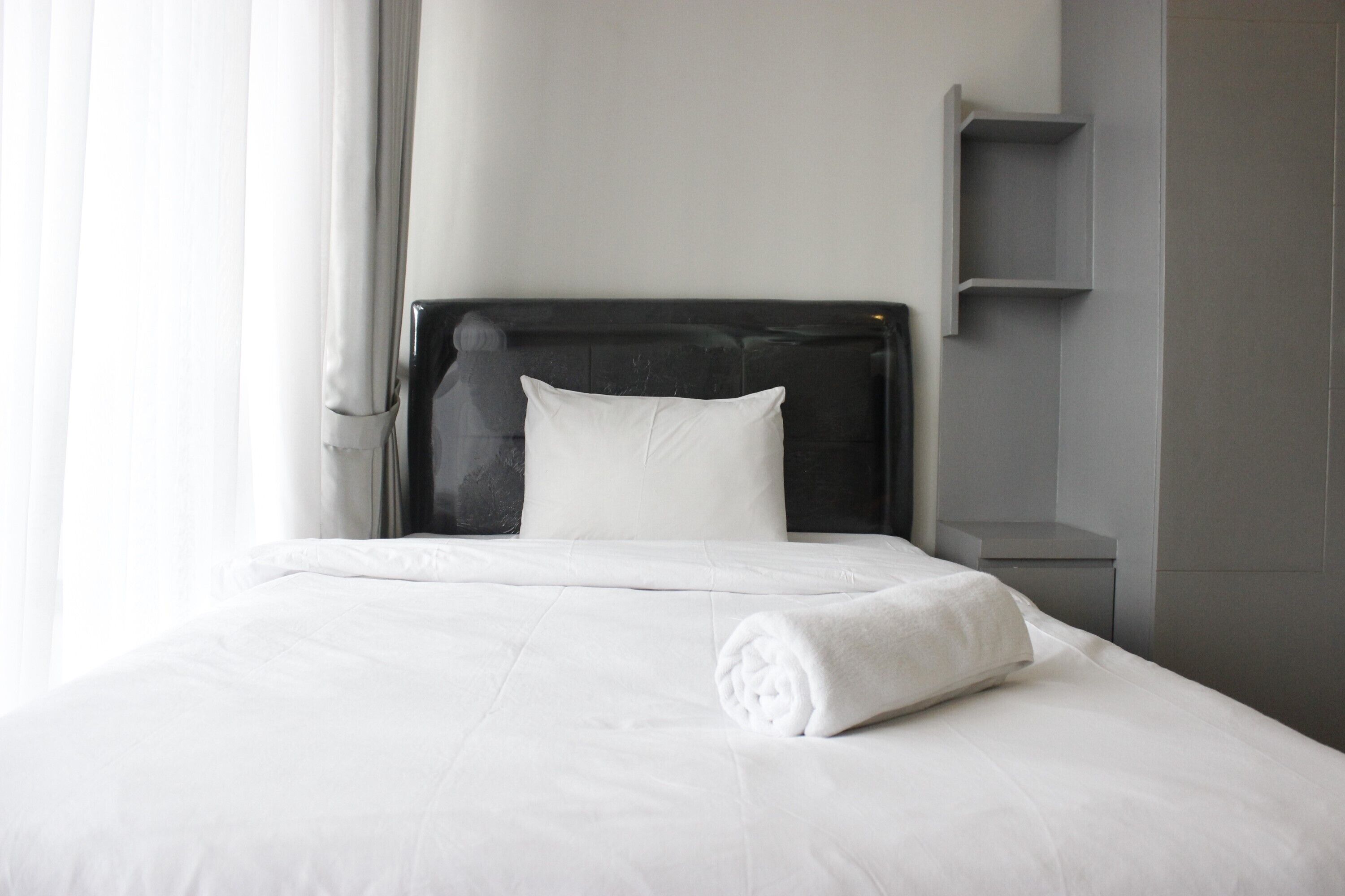 Una cama bien hecha con sábanas blancas y una toalla enrollada, un cabecero negro y una pared gris con una pequeña estantería.