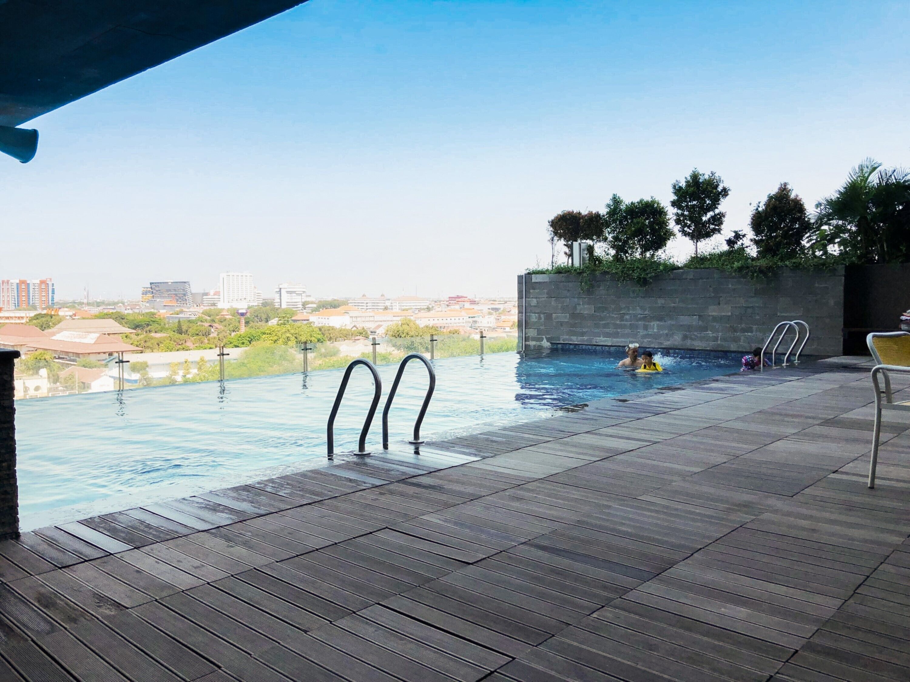 An infinity pool with a view of a cityscape, surrounded by a wooden deck and a stone wall.