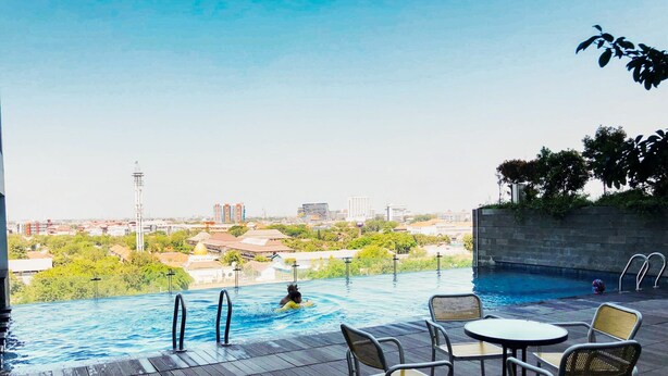 Una piscina exterior con terraza de madera, sillas de descanso y vistas a un paisaje urbano de fondo.