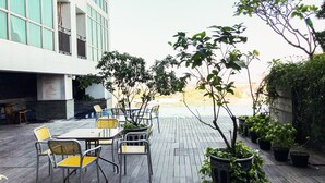 Un patio al aire libre con una mesa y sillas, rodeado de plantas en macetas y un edificio con grandes ventanales.
