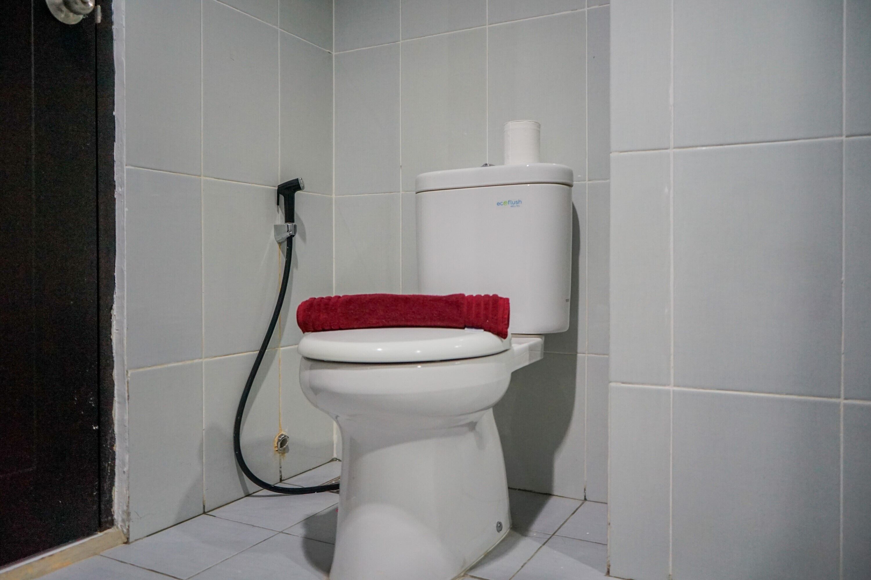 A white toilet with a red towel on the seat and a black hose attached to the wall.