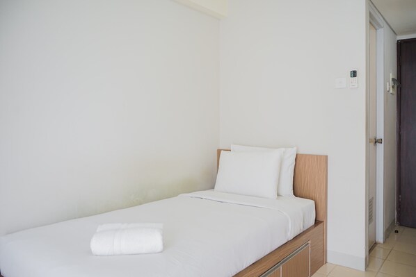 A small, neatly made bed with white linens and a folded towel, a wooden headboard, and a plain white wall.