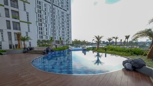 A rooftop swimming pool with a wooden deck, surrounded by palm trees and a high-rise building in the background. - Studio @ Akasa Pure Living Apartment BSD (South Tangerang)