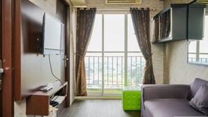 Hotellihuone, jossa on seinälle asennettu taulutelevisio, sohva, vihreä jalkarahi ja parveke, jossa on verhot.