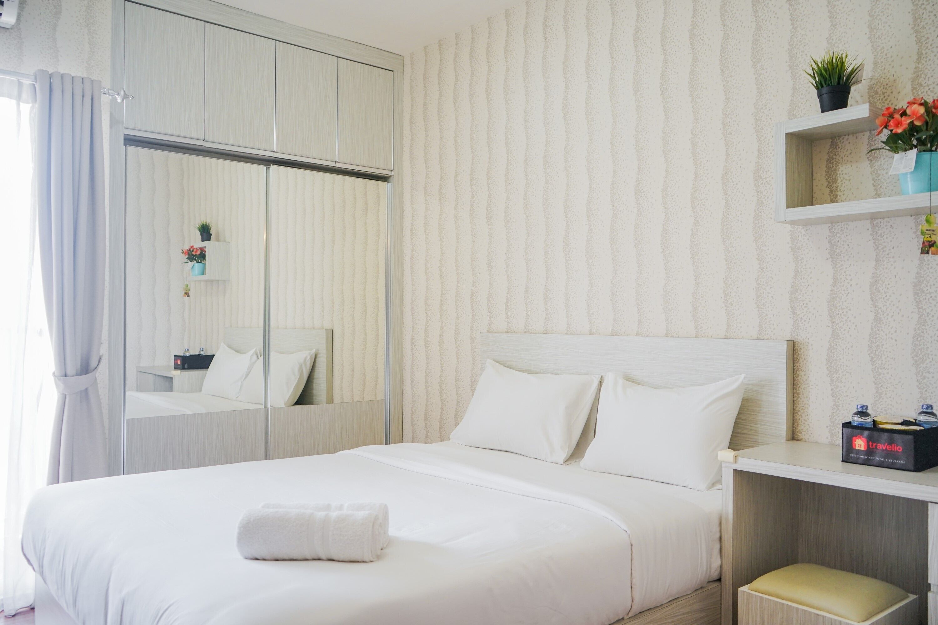 A neatly arranged hotel room with a large bed, a bedside table, a mirror, and a shelf with a plant and a box of toiletries.