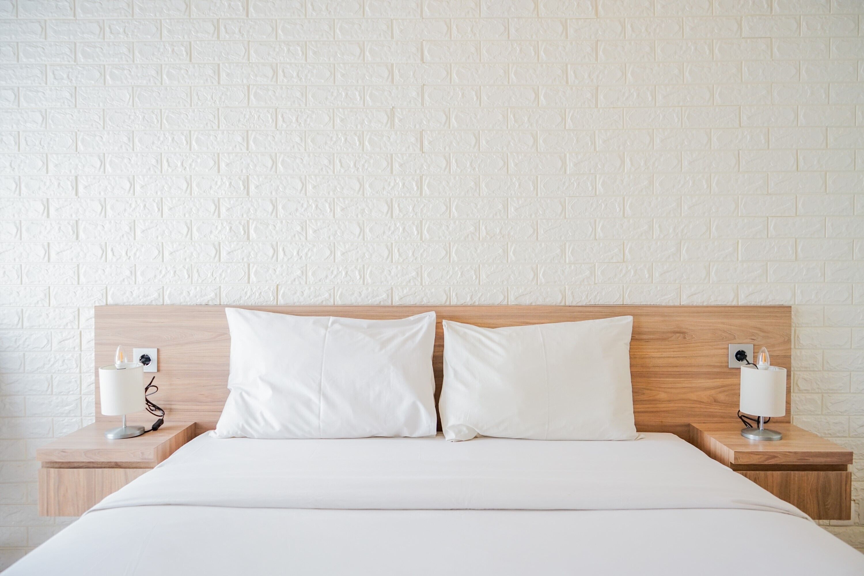 Un lit bien fait, avec du linge de lit blanc, deux tables de chevet avec des lampes et un mur décoré d’un motif en briques.