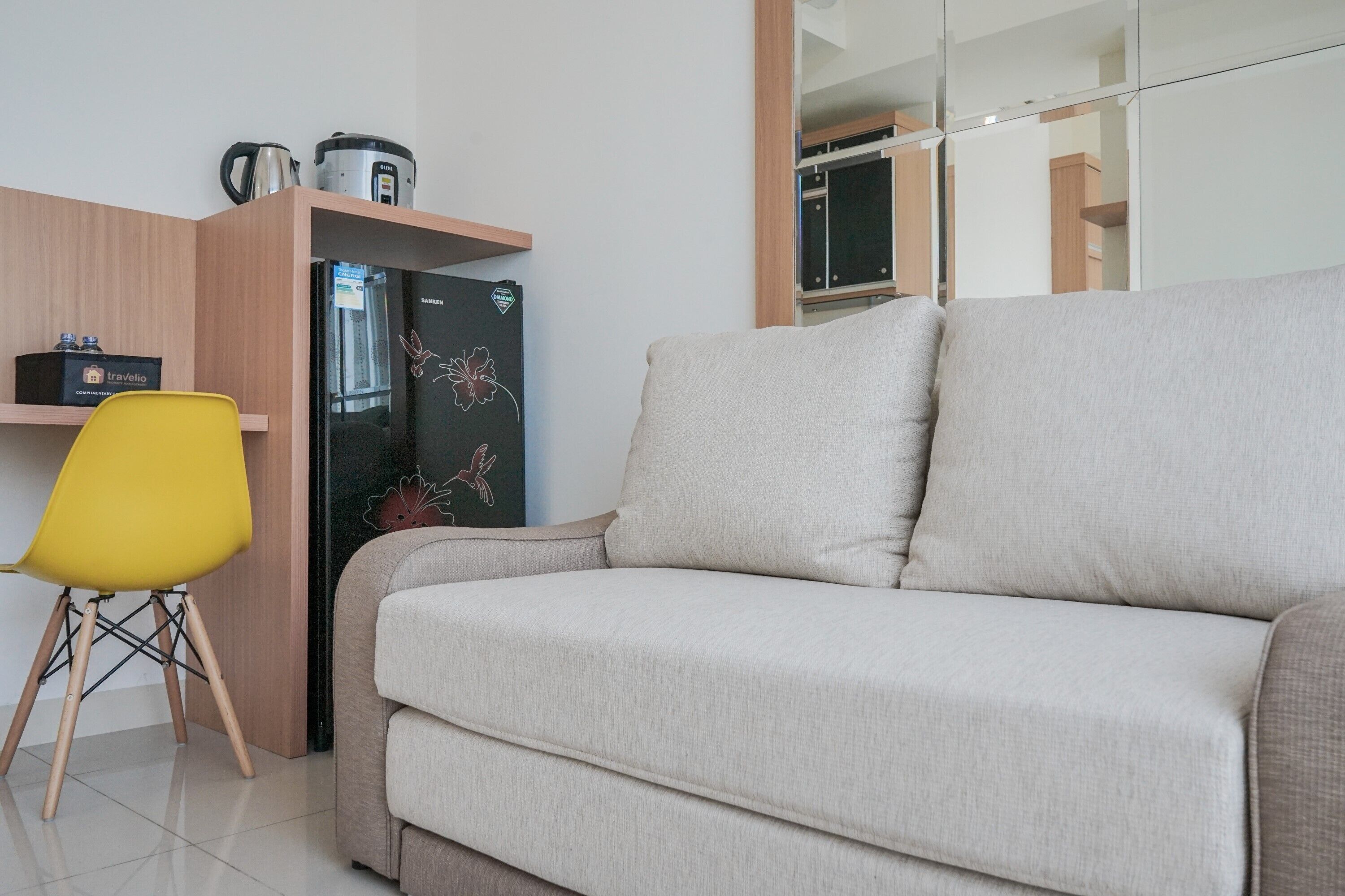 A modern living area with a beige sofa, a yellow chair, a black refrigerator with a logo, and a wooden shelf with kitchen items.