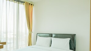 A neatly made bed with white linens and a single pillow, a wooden nightstand to the left, and a window with sheer curtains and a draped beige curtain. - Wonderful Studio Apartment at Tree Park (South Tangerang)