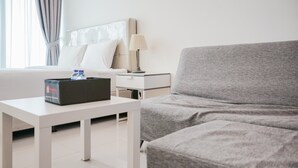A hotel room with a bed, a grey sofa, a white table with a laptop and water bottle, and a nightstand with a lamp. - Cozy and Relax Studio Room @ Tree Park BSD Apartment (South Tangerang)