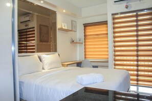 A hotel room with a bed, a desk, and a window with blinds. - Minimalist and Cozy Studio Room at Ayodhya Apartment (Tangerang)
