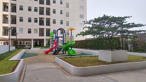Ein Spielplatz mit Rutschen und Klettergerüsten, umgeben von einer Grünanlage mit einem Baum und einem Gebäude im Hintergrund.