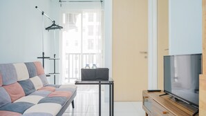 A modern living room with a sofa, a TV on a wooden stand, a table with a laptop, and a window with a view of buildings. - New Furnished 2BR near CGK Airport @ Ayodhya Apartment (Tangerang)