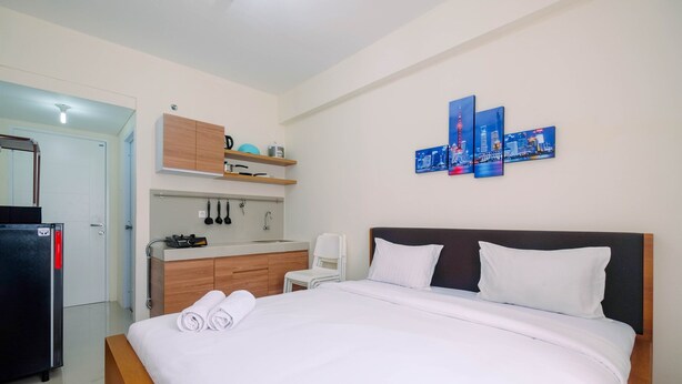 A hotel room with a bed, a small kitchenette, a refrigerator, a white chair, and a wall-mounted artwork.