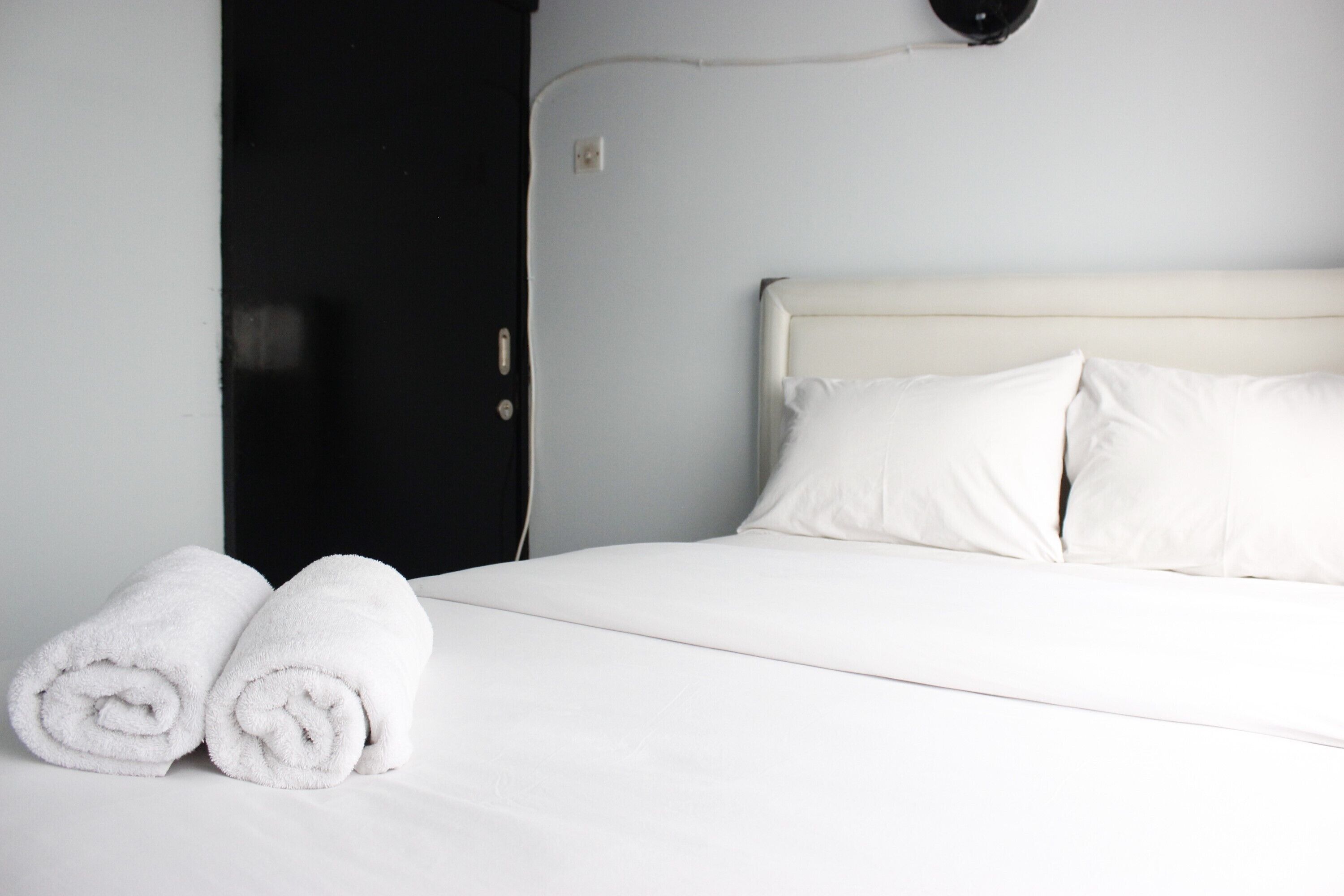 A neatly made bed with white linens and pillows, a black door, and a wall-mounted light fixture.