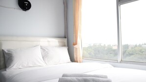 A neatly made bed with white linens and pillows, a window with a view of greenery, and a wall-mounted light fixture. - Quite 2BR Apartment at The Jarrdin Cihampelas (Bandung)