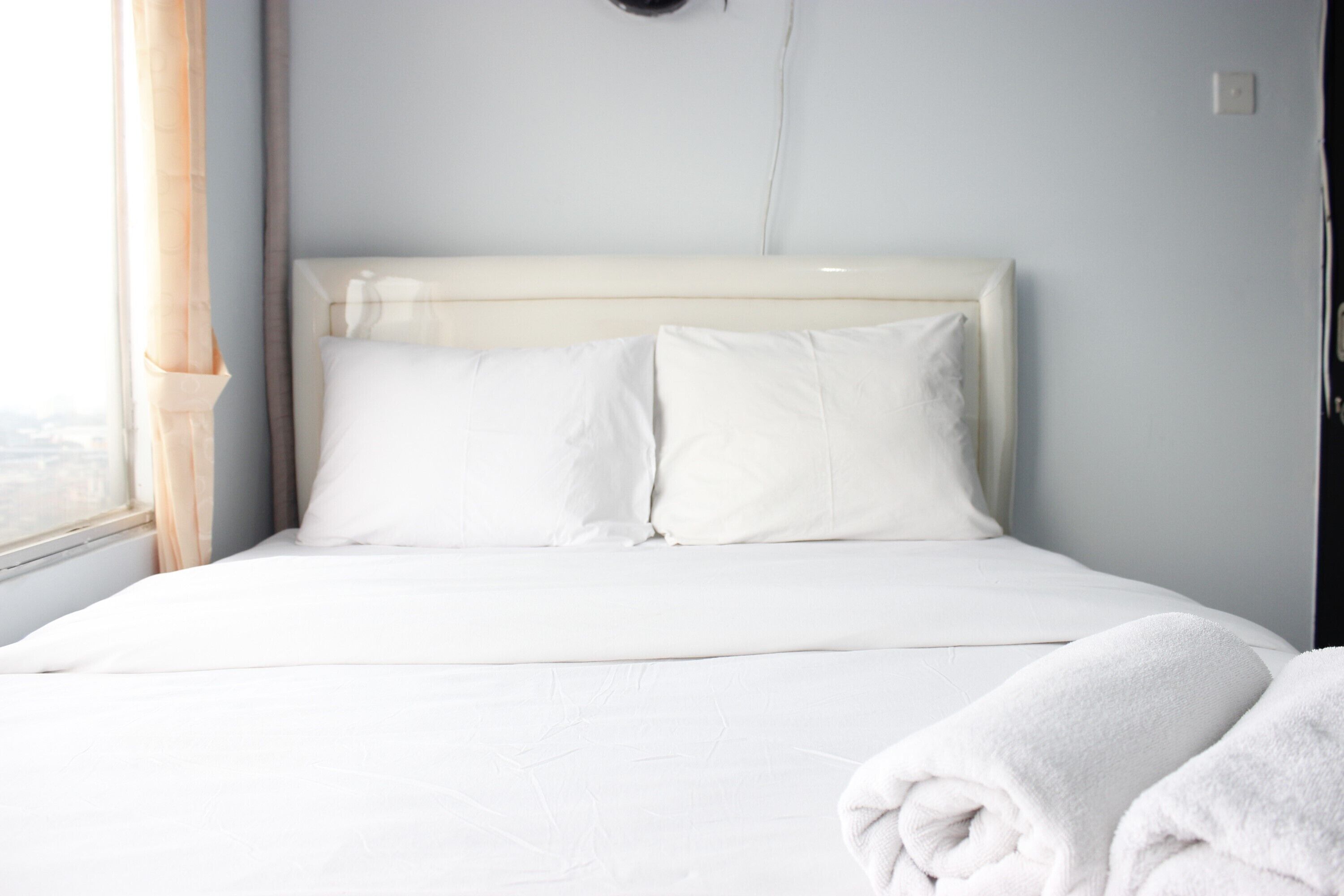 Uma cama bem arrumada com lençóis e almofadas brancas, uma janela com cortinas e uma cabeceira branca.