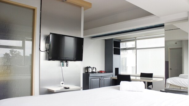 A modern hotel room with a large bed, a flat-screen TV mounted on the wall, a small table with a chair, and a window providing a view of the outdoors. - Spacious & Trendy Studio at Jarrdin Cihampelas Apartment (Bandung)