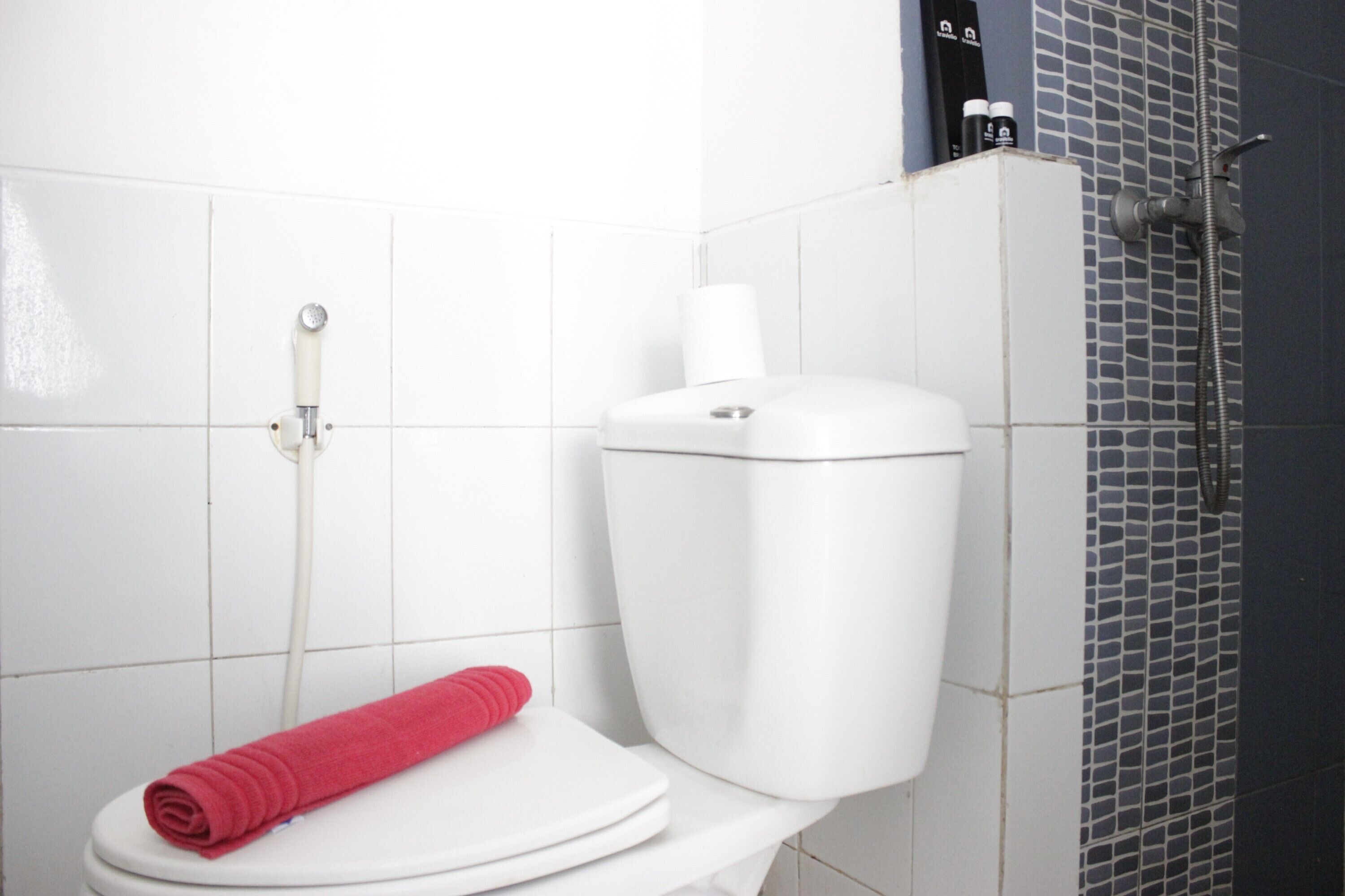 Et hvidt toilet med et rødt håndklæde, toiletpapirholder og brusehoved på væggen.