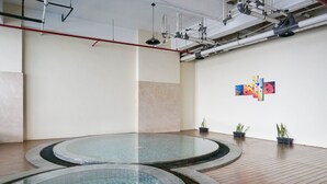 A modern indoor swimming pool with a wooden floor and a tiled edge. There are potted plants on the wall and a ceiling with visible pipes and lighting fixtures.