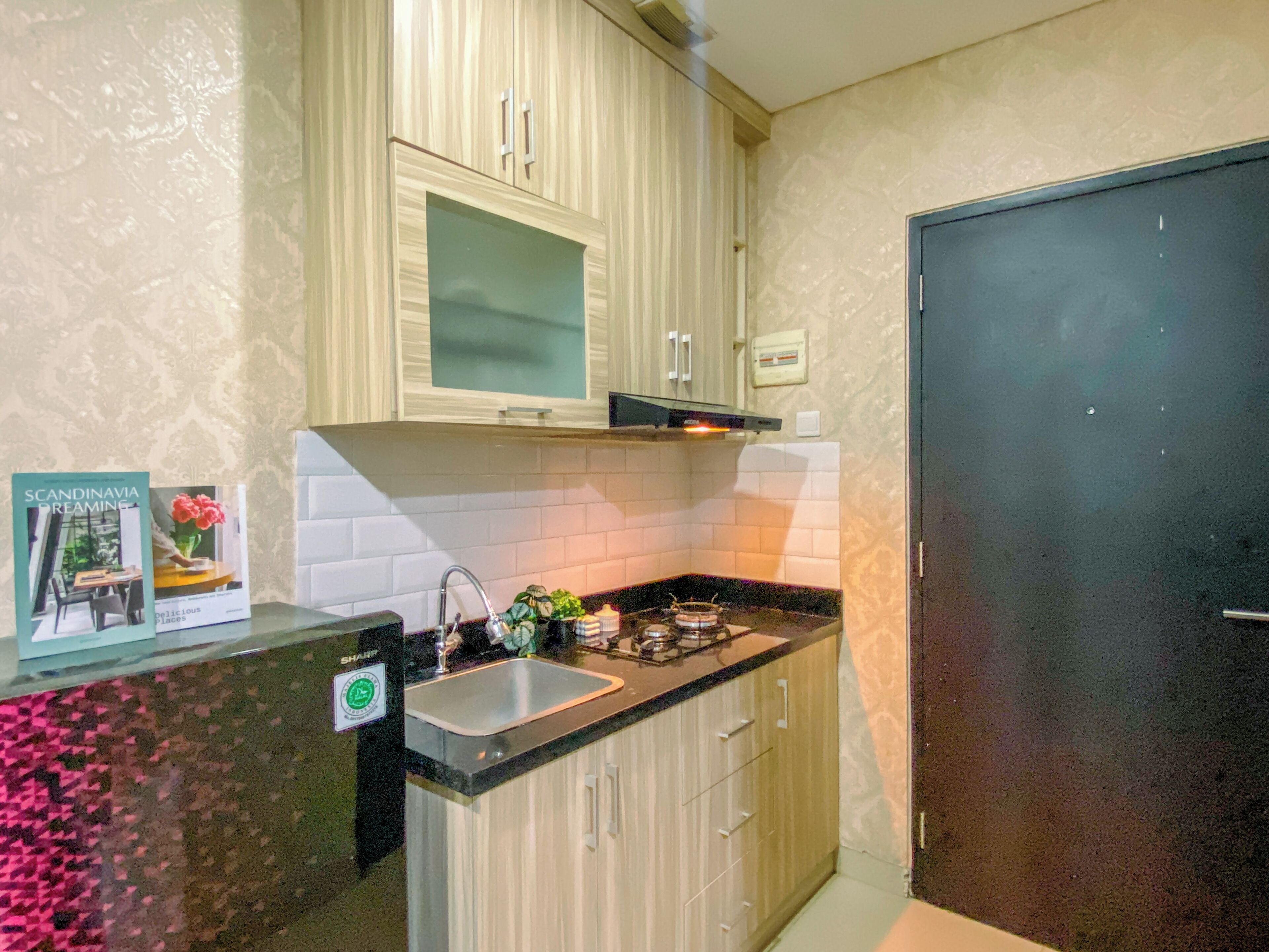 A kitchen with light wood cabinets, a stainless steel sink, and a black countertop. There is a stove with a pot on it, and a small plant on the counter. A door is visible on the right side of the image.