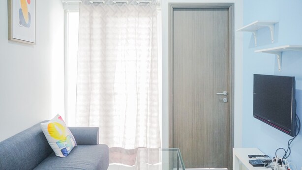 A living room with a grey sofa, a glass coffee table, a TV mounted on the wall, a door, and a window with sheer curtains. - Comfy and Stylish 2BR Emerald Bintaro Apartment (South Tangerang)