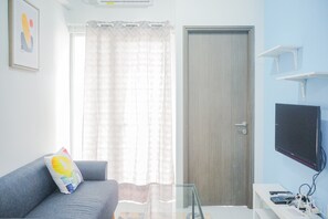 A living room with a grey sofa, a glass coffee table, a TV mounted on the wall, a door, and a window with sheer curtains. - Comfy and Stylish 2BR Emerald Bintaro Apartment (South Tangerang)