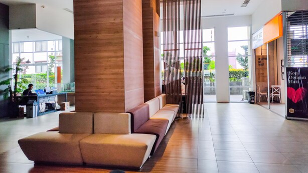 A modern lobby with a seating area, a reception desk, and a coffee shop. - Simply Look Studio Tamansari Mahogany Karawang Apartment (Karawang)