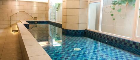 A swimming pool with blue mosaic tiles, a tiled deck, and a glass wall with plants.
