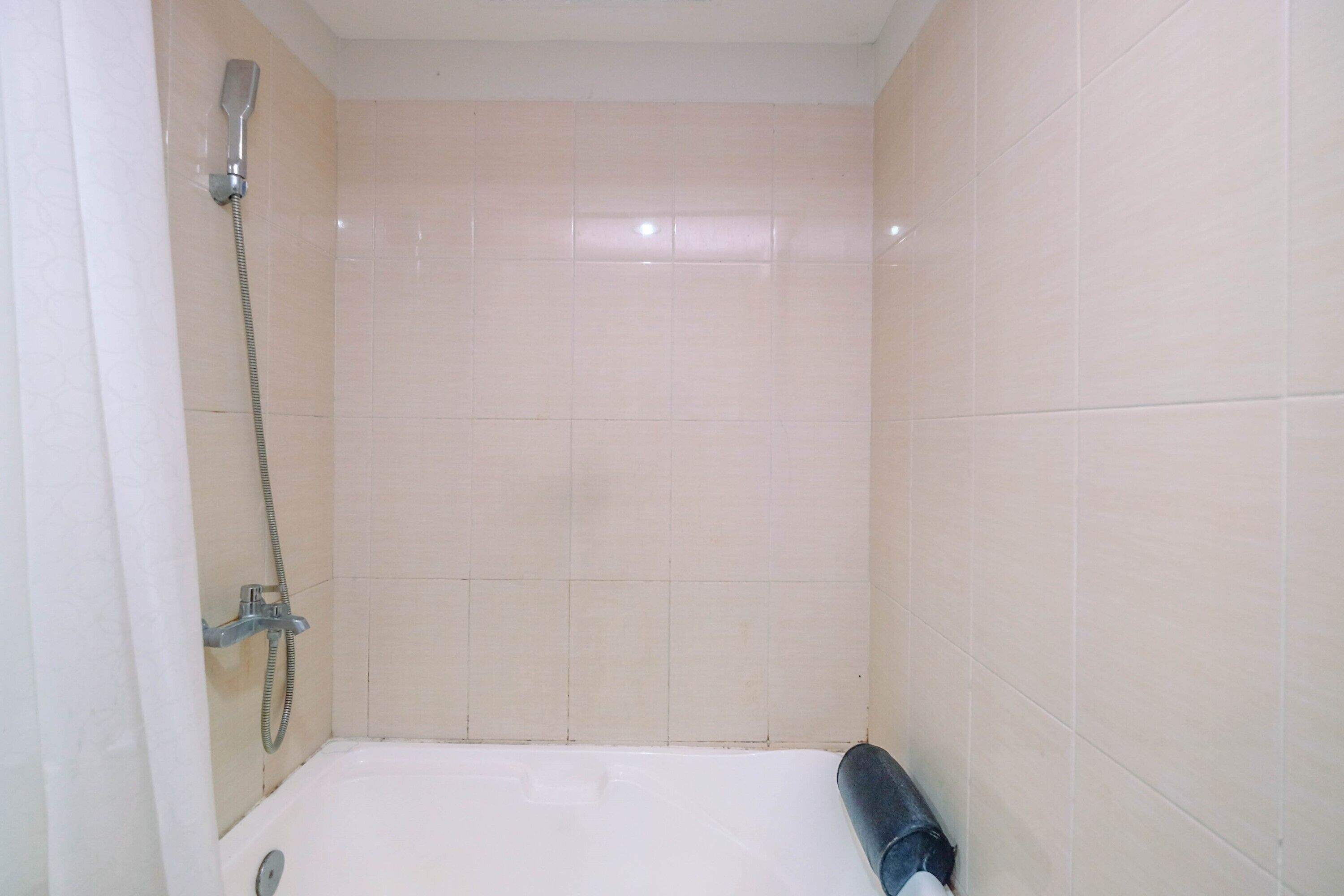 A small, tiled shower room with a handheld showerhead and a roll of toilet paper on the floor.