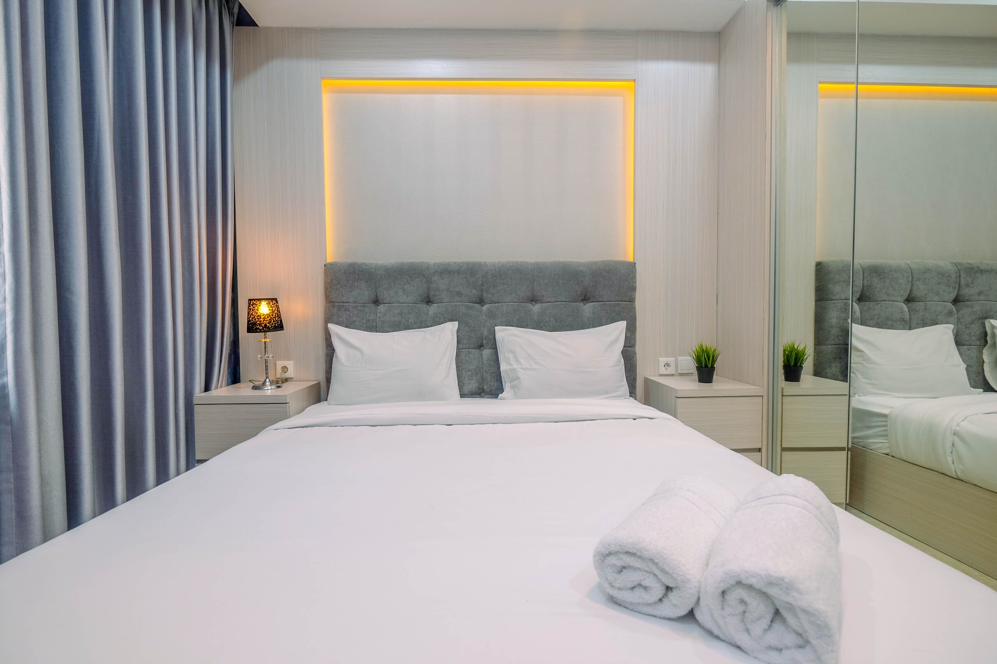A neatly made bed with white linens and two rolled towels, a bedside lamp, and a small potted plant on a nightstand.