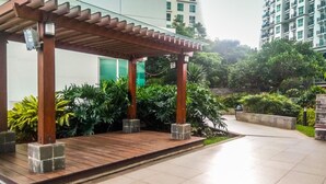 A covered outdoor area with wooden pillars, surrounded by greenery and a tiled floor.