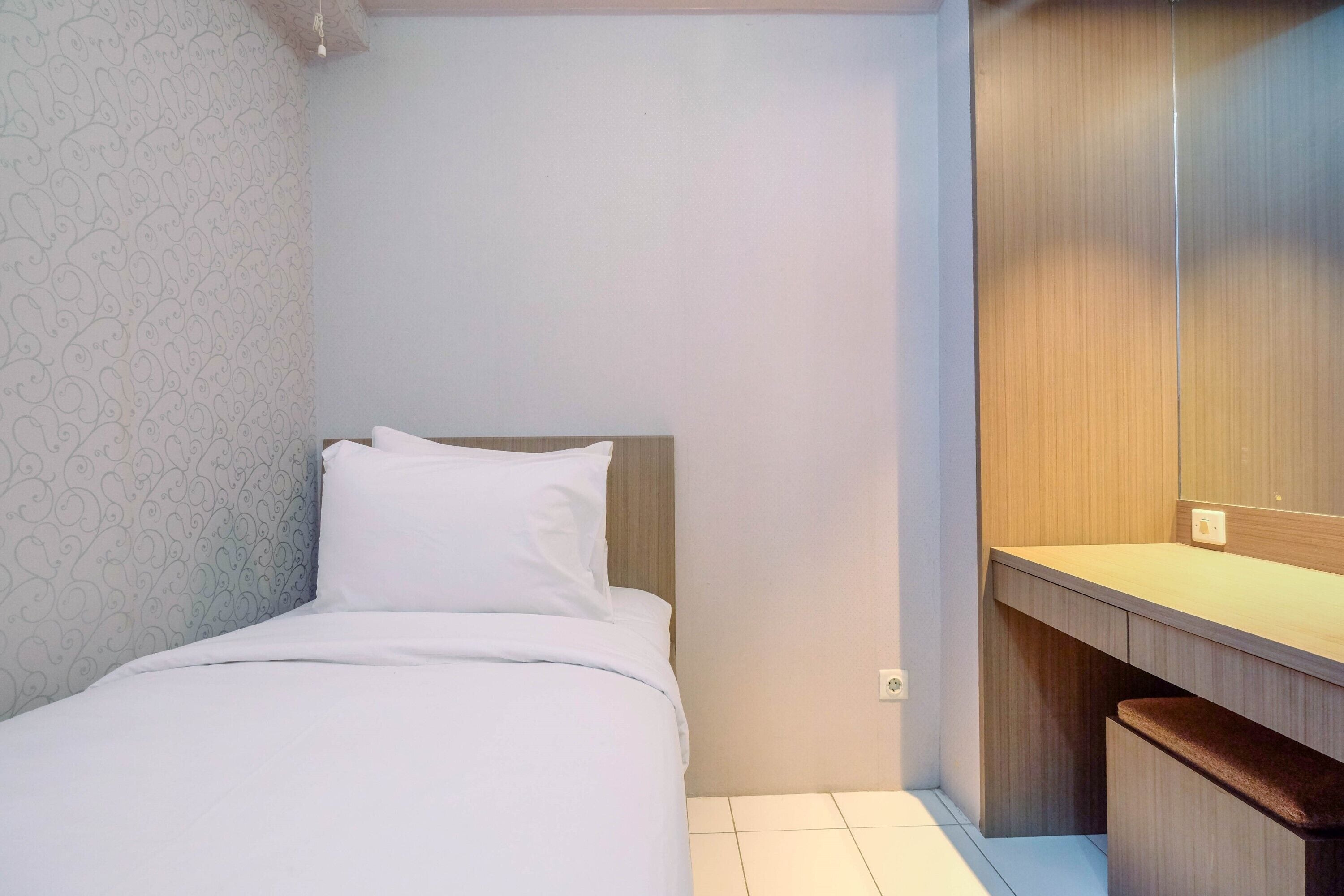 Un petit lit bien fait, avec une literie blanche, dans une chambre compacte dotée d’une tête de lit en bois et d’un petit banc en bois.