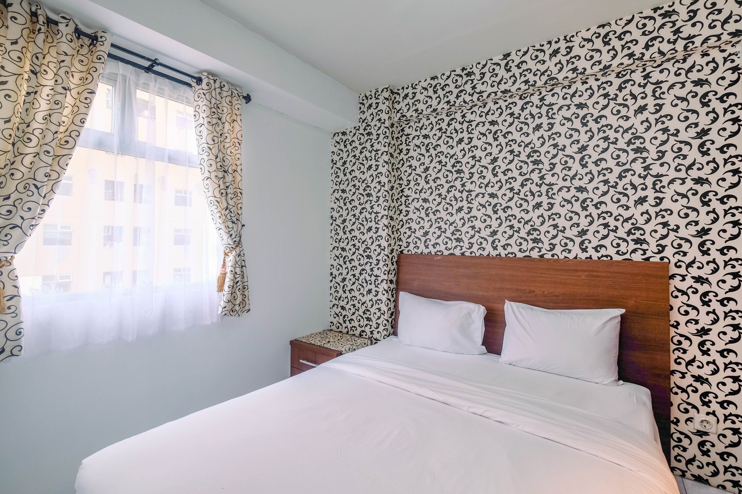 A neatly made bed with white linens, a wooden headboard, and a nightstand with a small drawer. The room features patterned wallpaper and sheer curtains on a window.