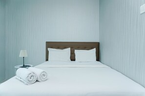 A neatly made bed with white linens, a wooden headboard, and two pillows. There is a small bedside table with a lamp and rolled towels. - New Furnished and Comfortable Studio Woodland Park Apartment (Jakarta)