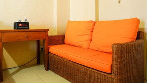 A wicker sofa with orange cushions, a wooden table with a travelio box, and a wall-mounted phone. - Comfort Stay Studio Room @ Green Palace Kalibata Apartment (Jakarta)