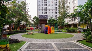 A playground with colorful play structures surrounded by a landscaped garden, a circular paved area, and a tall residential building in the background. - Wonderful and Cozy Studio at Green Palace Apartment (Jakarta)