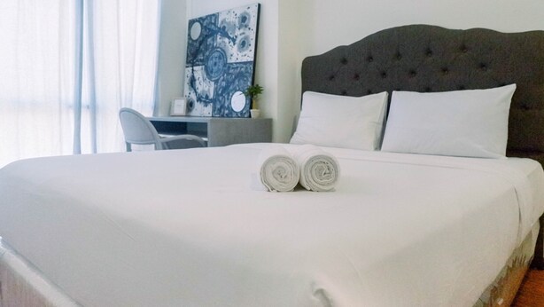 Una cama bien tendida con sábanas blancas y dos toallas enrolladas, un cabecero capitonado y un cuadro en la pared.