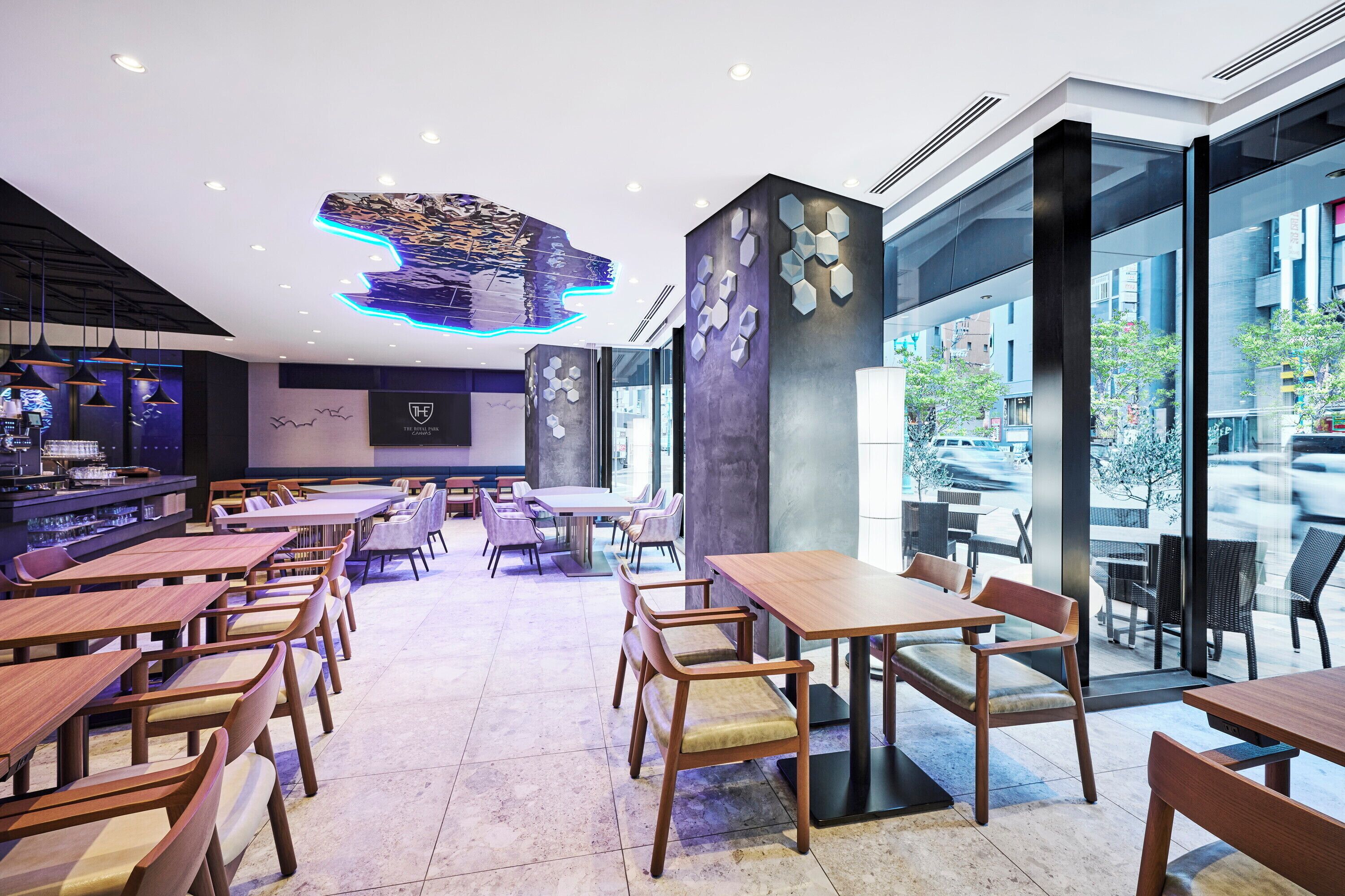 A modern restaurant with wooden tables and chairs, a bar area, and large windows offering a view of the street.